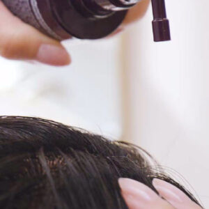 A person applies Invati Advanced Scalp Revitalizer to their scalp using a dark-colored pump dispenser, while another hand touches the hair near the application area. - SAAB Salon Spa | Ottawa On, Canada