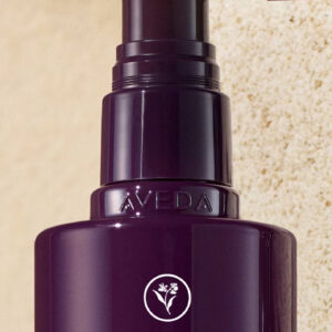 Close-up of a dark purple Invati advanced scalp revitalizer pump bottle against a beige textured background with text stating, 