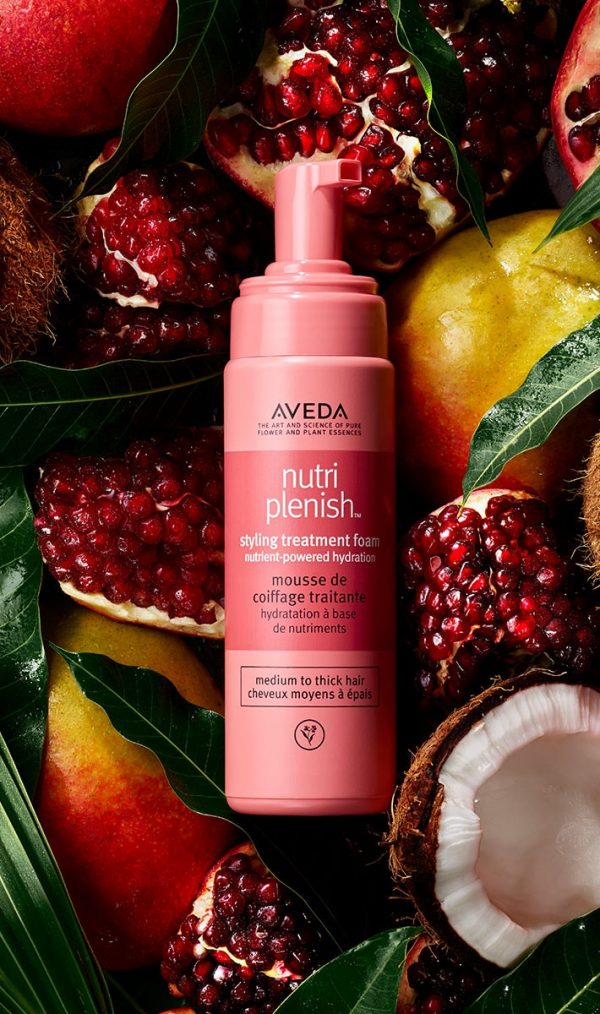 A pink bottle of Nutriplenish Styling Treatment Foam by Aveda is surrounded by pomegranates, leaves, and a coconut half. Ideal for medium to thick hair, it complements beautiful skin and enhances your natural hair color. - SAAB Salon Spa | Ottawa On, Canada