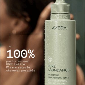 A green bottle of Pure Abundance Volumizing Conditioning Rinse made from 100% post-consumer HDPE stands in focus, with a blurred person in the background. - SAAB Salon Spa | Ottawa On, Canada