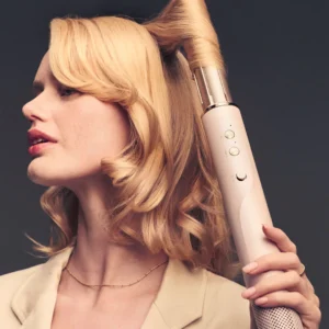 A blonde woman curls her hair with the T3 Aire360, a large cylindrical automatic curling iron, against a dark background. - SAAB Salon Spa | Ottawa On, Canada