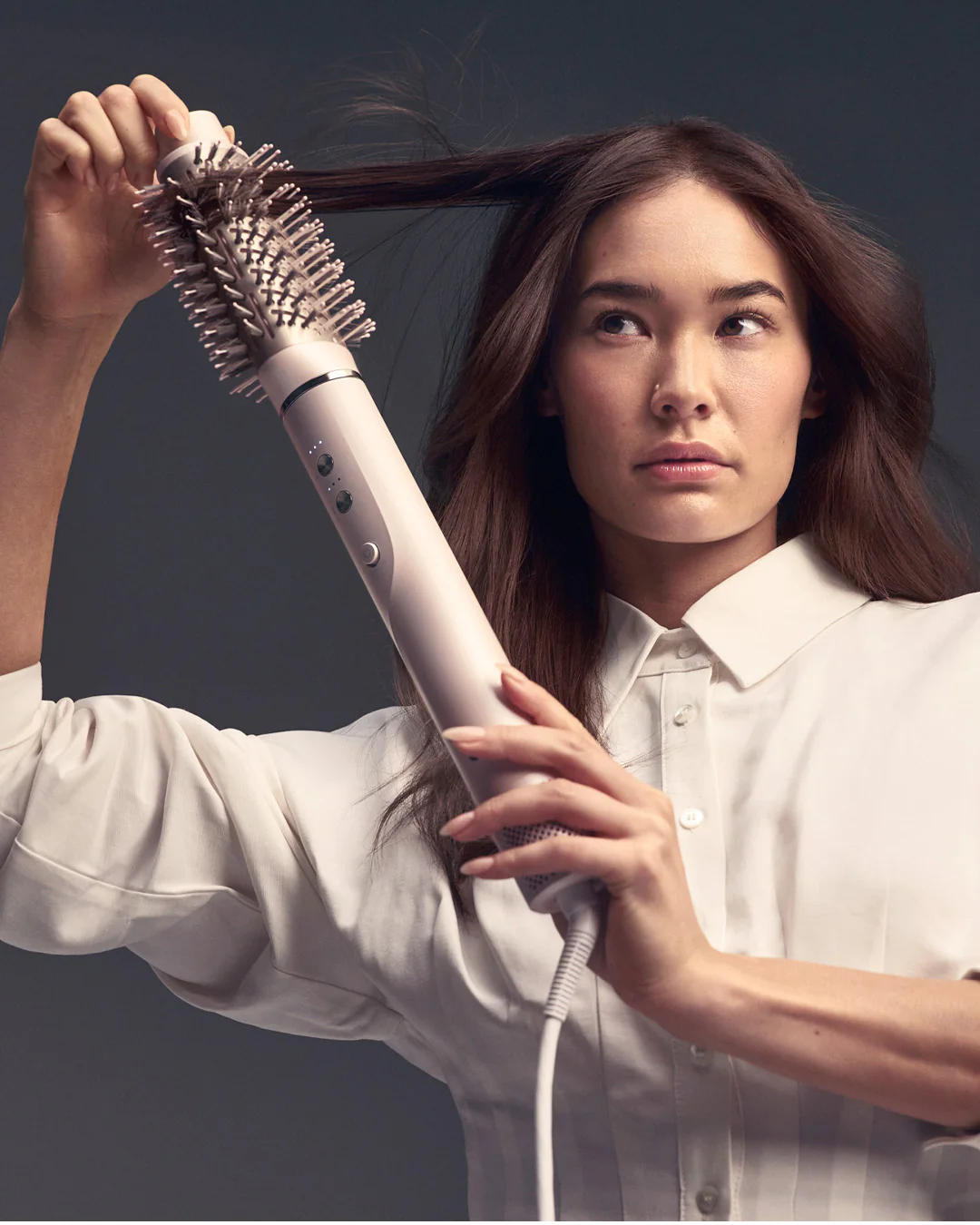 A woman in a white shirt styles her long brown hair with the T3 Aire360 electric round brush against a neutral background. - SAAB Salon Spa | Ottawa On, Canada